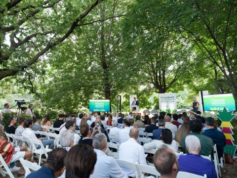 Pubblico seduto a un evento all’aperto in un parco, con palco e cartelli verdi