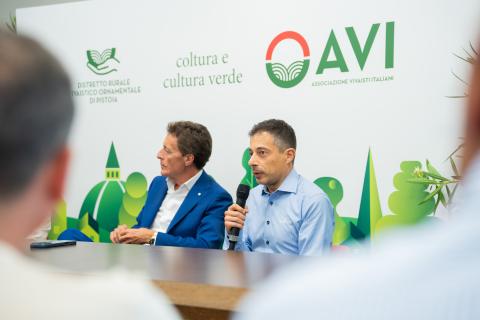 Due uomini in conferenza stampa, uno parla al microfono davanti a un logo OAVI