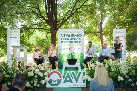 Conferenza all’aperto con relatori su palco e cartello “VIVAISMO LAGHETTI”