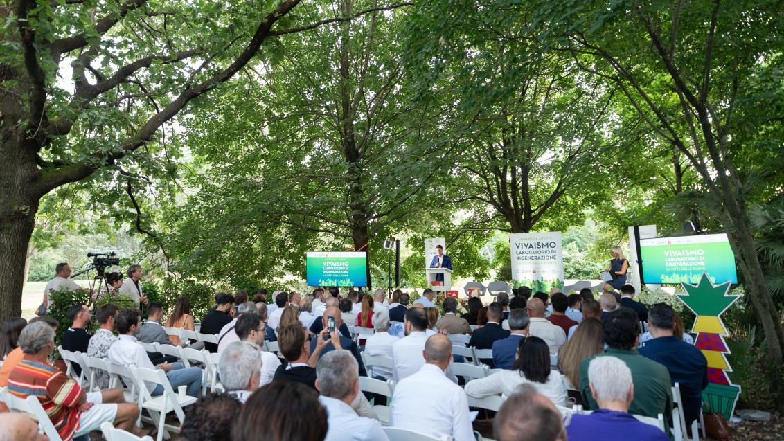 Pubblico seduto in un parco, ascolta un evento con pannelli verdi sullo sfondo