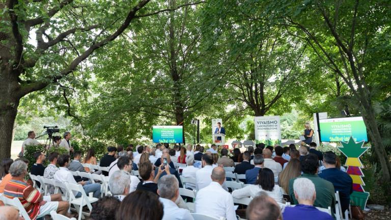 Pubblico seduto a un evento all’aperto in un parco, con palco e cartelli verdi