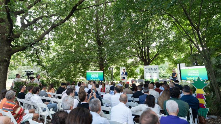 Pubblico seduto in un parco, ascolta un evento con pannelli verdi sullo sfondo